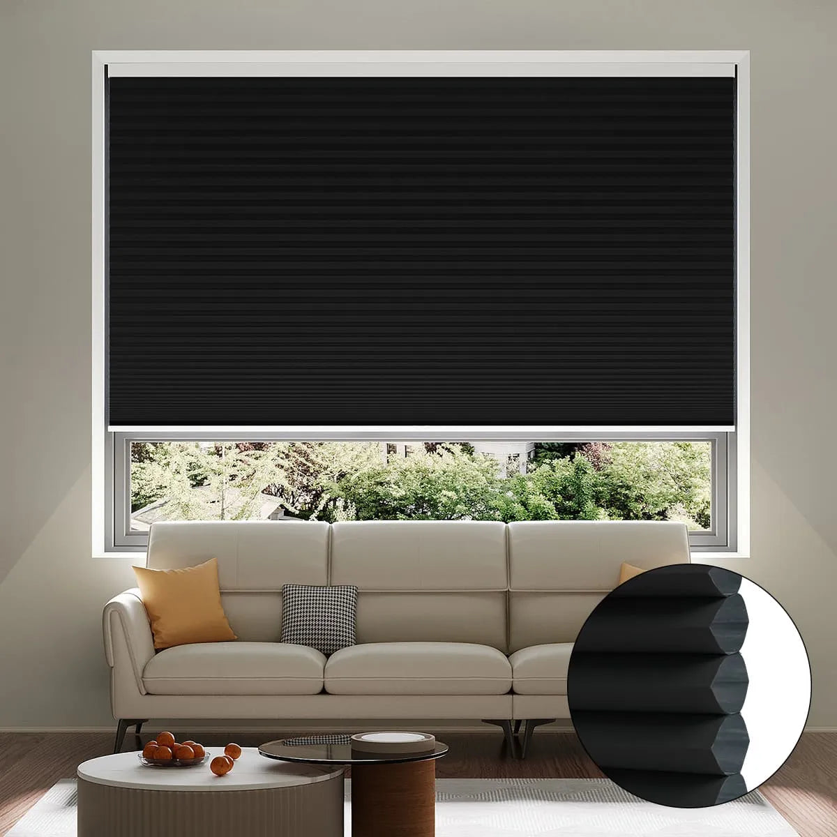 A living room with a beige sofa, yellow and orange cushions, a coffee table, and a large window covered by a closed black cellular blackout shade; an inset shows a close-up of the shade’s honeycomb structure.