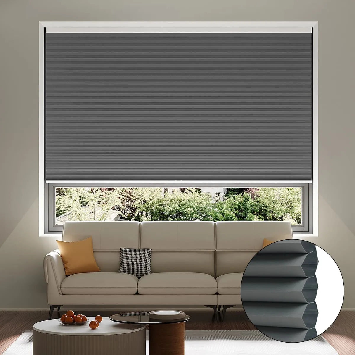 A living room with a white sofa, cushions, and a coffee table. The window features a partially lowered dark grey honeycomb cellular blind, with an inset showing the blind’s layered structure. Trees are visible outside.