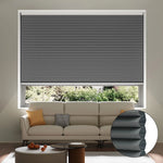 A living room with a white sofa, cushions, and a coffee table. The window features a partially lowered dark grey honeycomb cellular blind, with an inset showing the blind’s layered structure. Trees are visible outside.