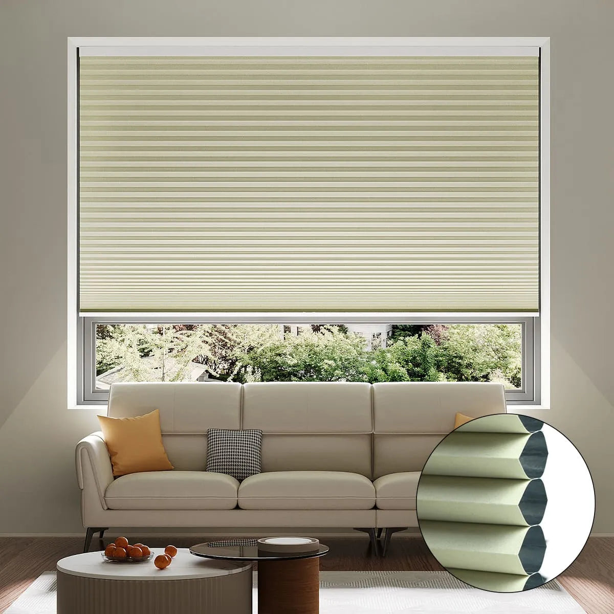 A living room with a beige pleated cellular window shade, cream sofa with yellow and gray pillows, a small coffee table, and a window view of greenery outside. An inset shows a close-up of the shade’s honeycomb structure.
